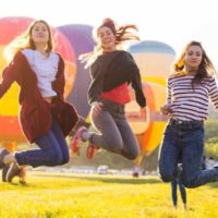 Três raparigas a saltar num campo verde com balões de ar quente ao fundo