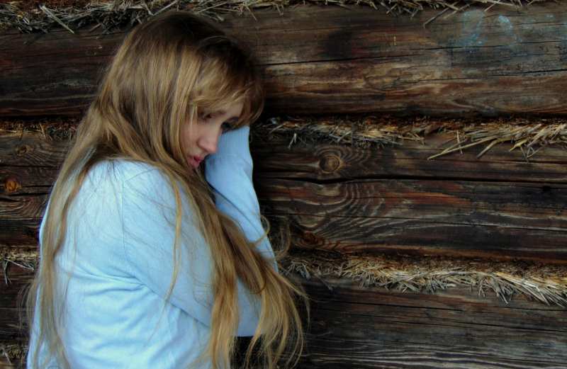 Mulher loira triste com cabelo comprido sentada junto a um muro de madeira 