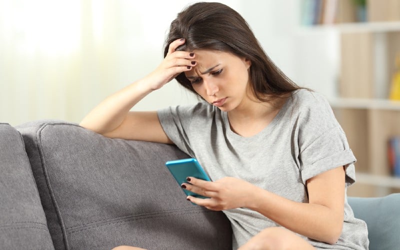 Chica triste sentada en un sofá sosteniendo un smartphone en una mano