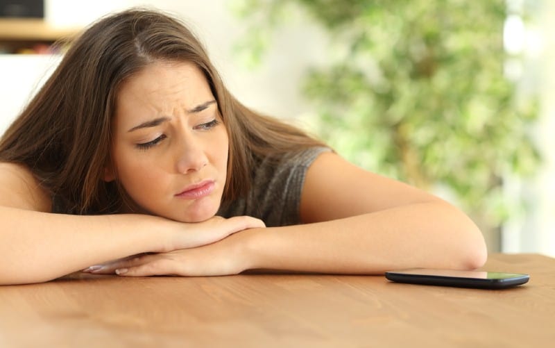 sad girl with hands on a table and head on her hands looking at a phone on the table