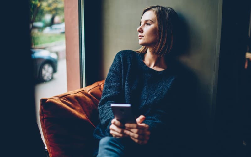 Mujer triste sentada cerca de una ventana con el móvil en una mano
