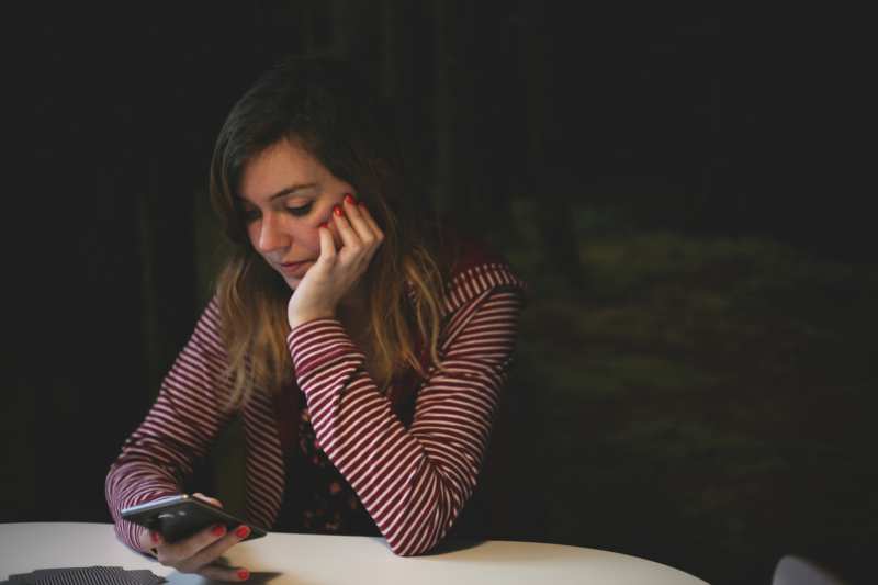 Joven pensativa enviando mensajes de texto en su teléfono mientras está sentada en una mesa