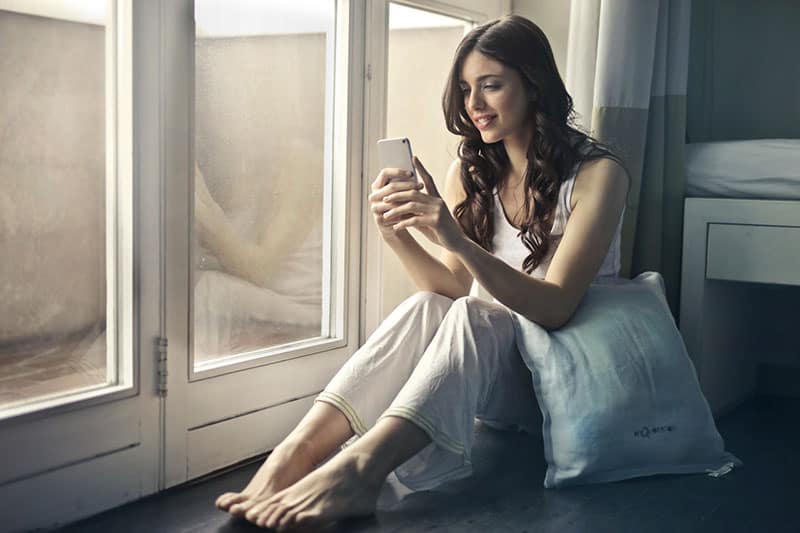 woman in white holding cellphone and a pillow beside her