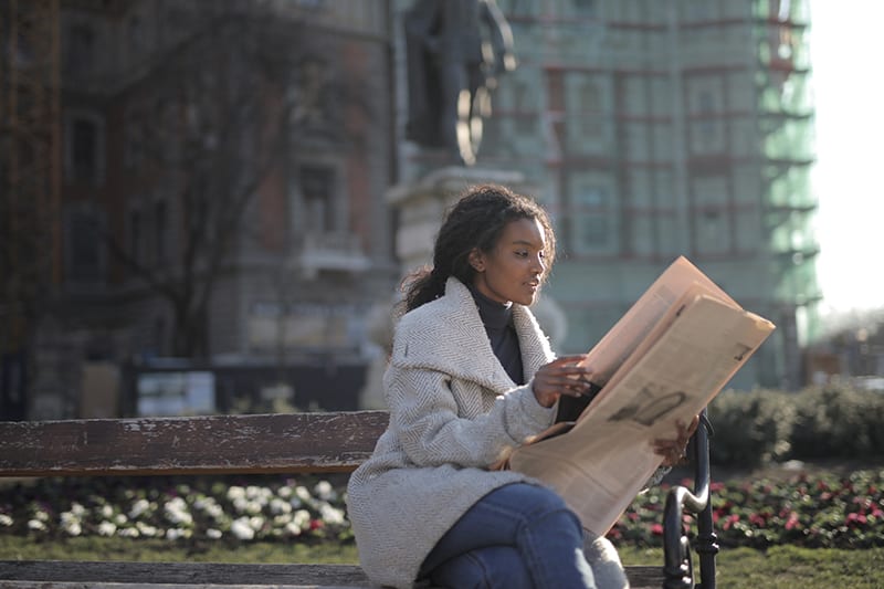 woman in gray coat sitting on the bench and reading newspaper