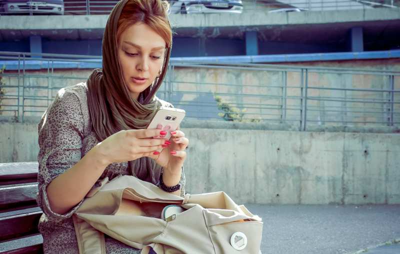 Mujer con pañuelo en la cabeza sentada en un banco y escribiendo mensajes en su teléfono