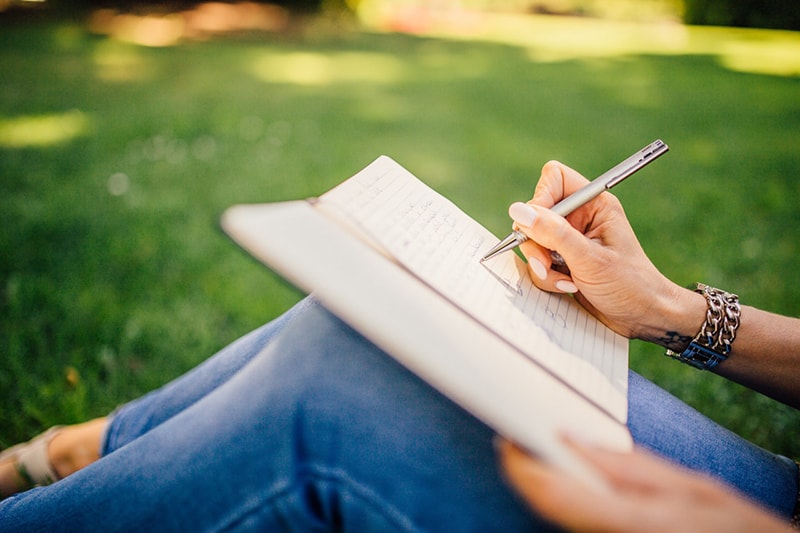 woman writing in the notebook while she is sitting on the grass in the park