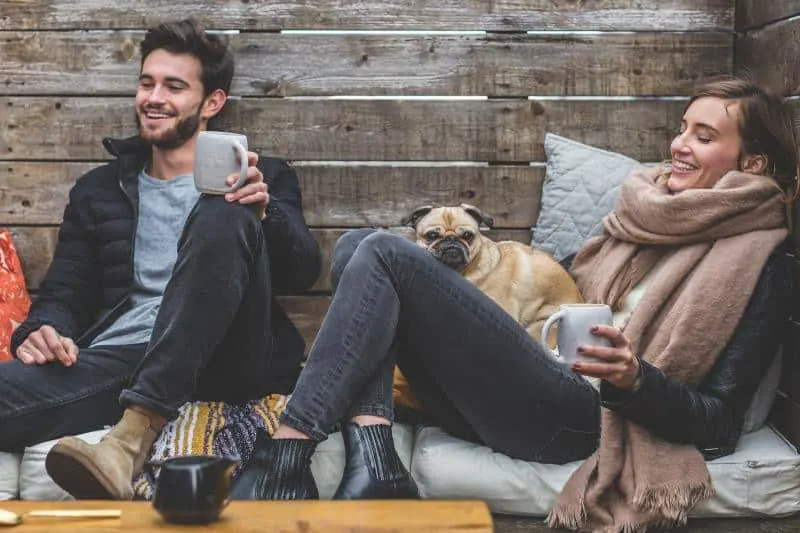 Young smiling couple with dog and tea cups sitting on a mattress by a wood wall