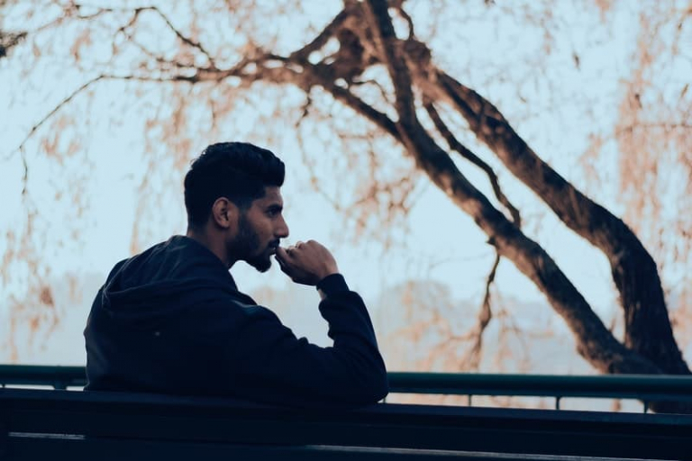 sideview of a man sitting on a bench near a bark of a tree with thumb on his mouth