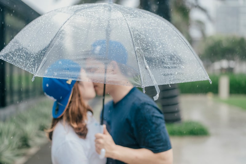 casal a beijar-se debaixo de um guarda-chuva, ambos com bonés azuis