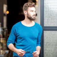 homem de camisola azul com um óculo na mão, parado à porta