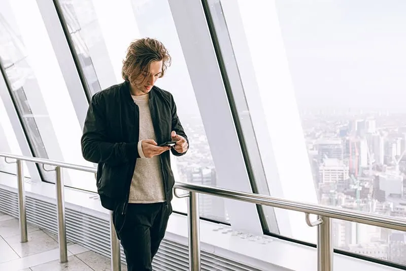 Man texting on phone wearing black jacket beside large window