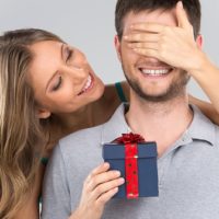 Woman covers man's eyes holding a gift box in one hand