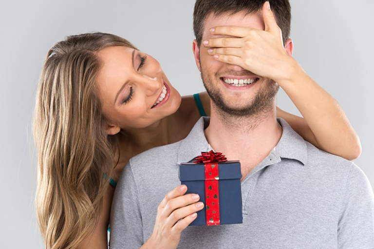 Woman covers man's eyes holding a gift box in one hand