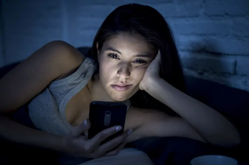 Woman propped up on bed looking bored with phone light hitting her face