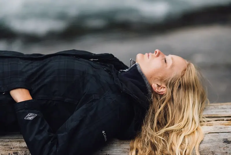 Woman wearing black jacket lies down on a large log by the beach