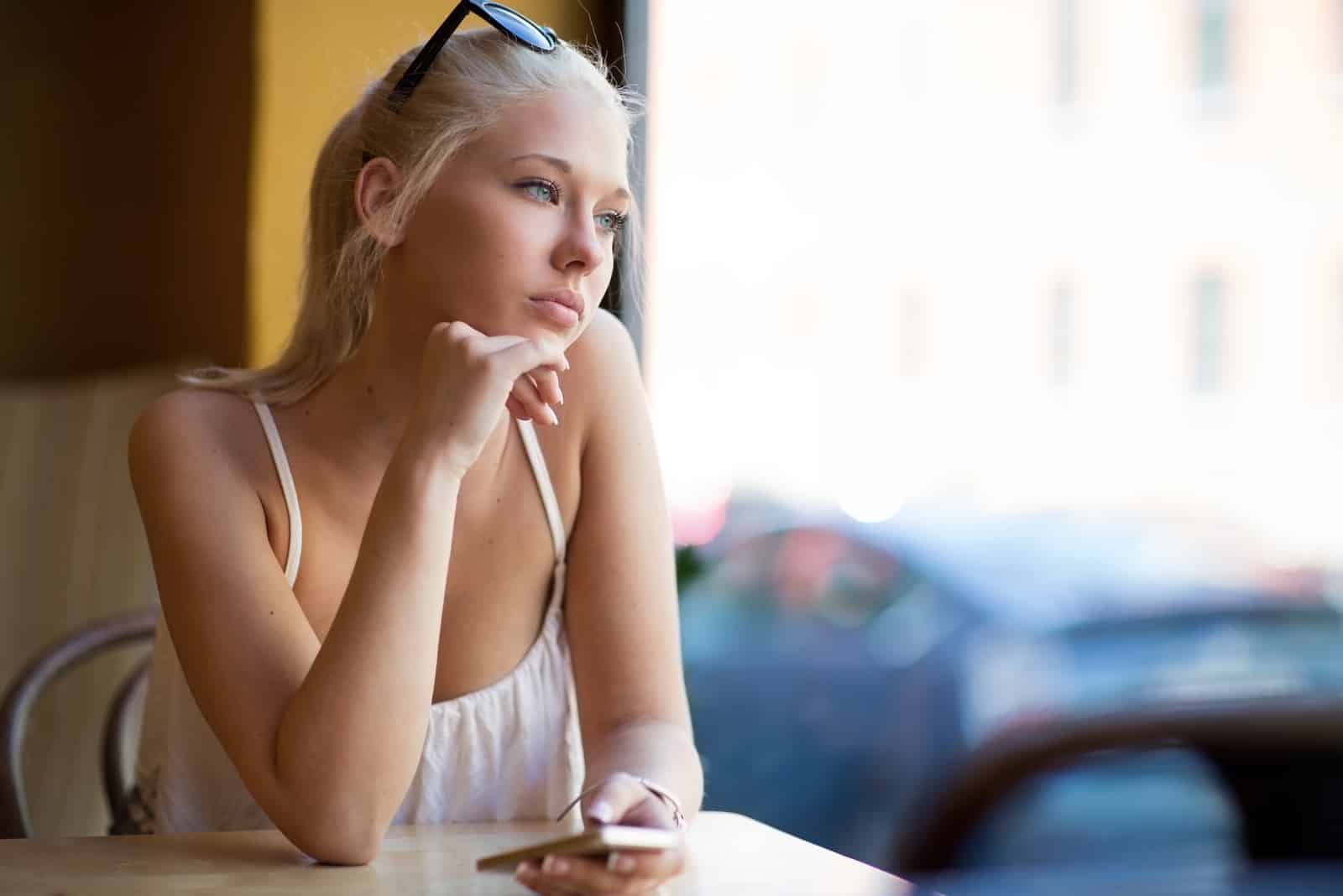 a beautiful woman with blonde hair tied in a ponytail sits thoughtfully at the table