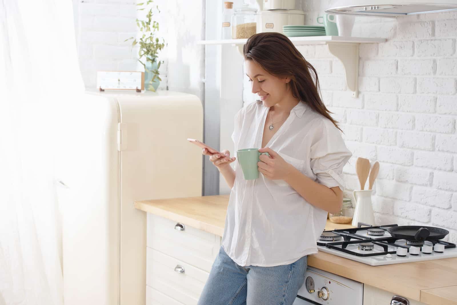 a smiling woman with a cup of coffee standing in the kitchen and pressing a phone