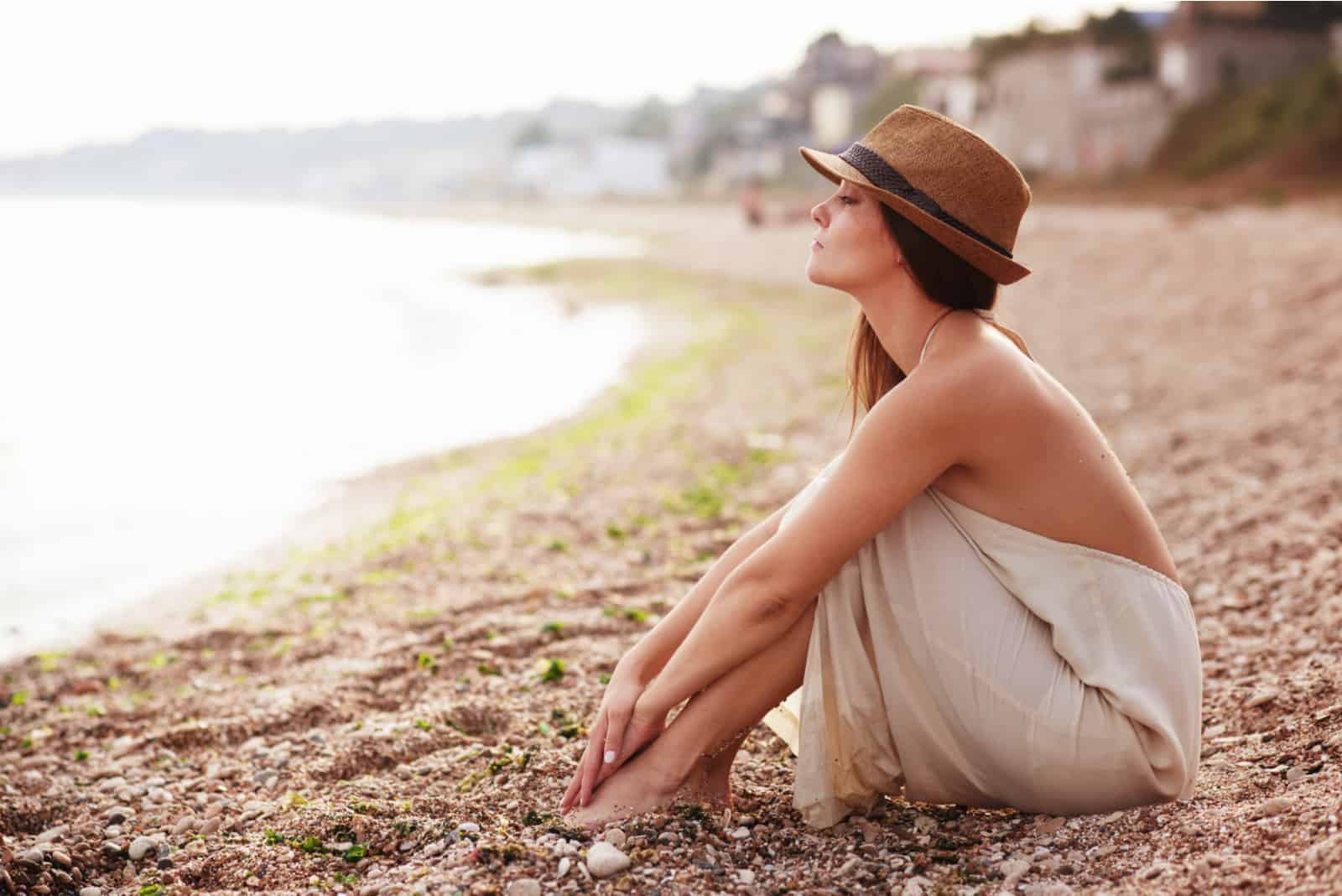 a woman with a hat on her head sits pensively on the beach and looks out to sea