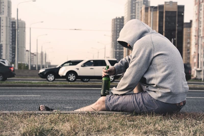 alcoholic man in street holding beer beside the street wearing a shirt with hood on