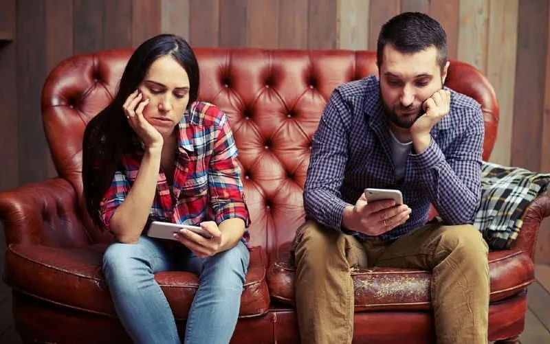 Bored couple looking at phones sitting on red couch