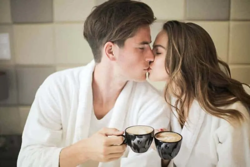 couple in white bathrobes kissing while holding black ceramic cups of coffee