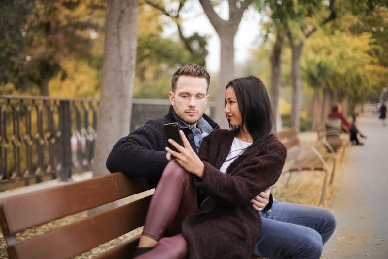 man and woman sitting on bench while looking at phone
