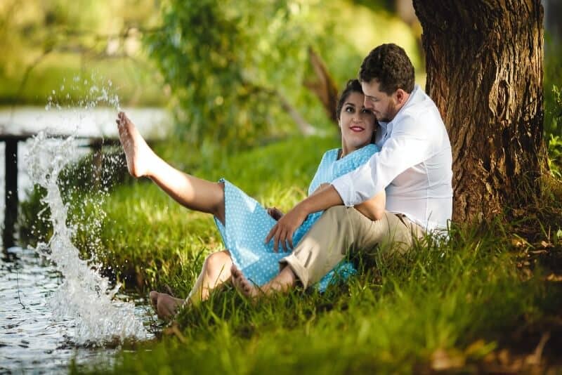 hombre y mujer sentados en la hierba bajo un árbol junto al río