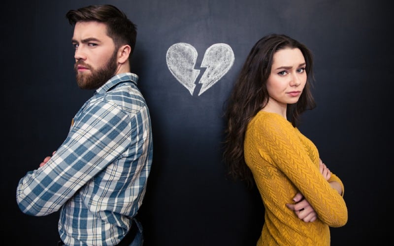 Jovem casal infeliz de pé, de braços cruzados, tendo como fundo um quadro de giz com um coração partido desenhado