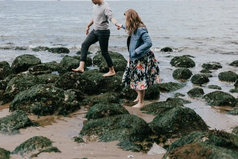 man and woman holding hands while stepping on rocks near sea