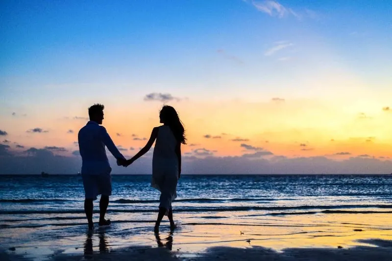 man and woman holding hands walking on seashore during golden hour