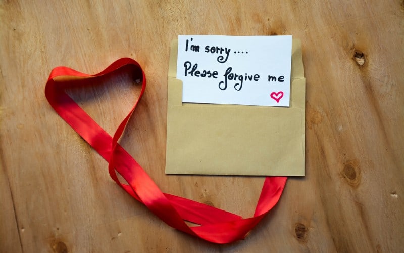 Heart made of red ribbon and an envelope with i'm sorry please forgive me text on a note on rustic wooden background