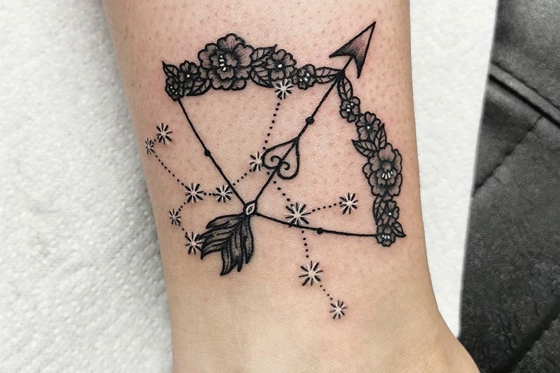 tatuagem de arco floral, flecha com penas e constelação de sagitário no tornozelo