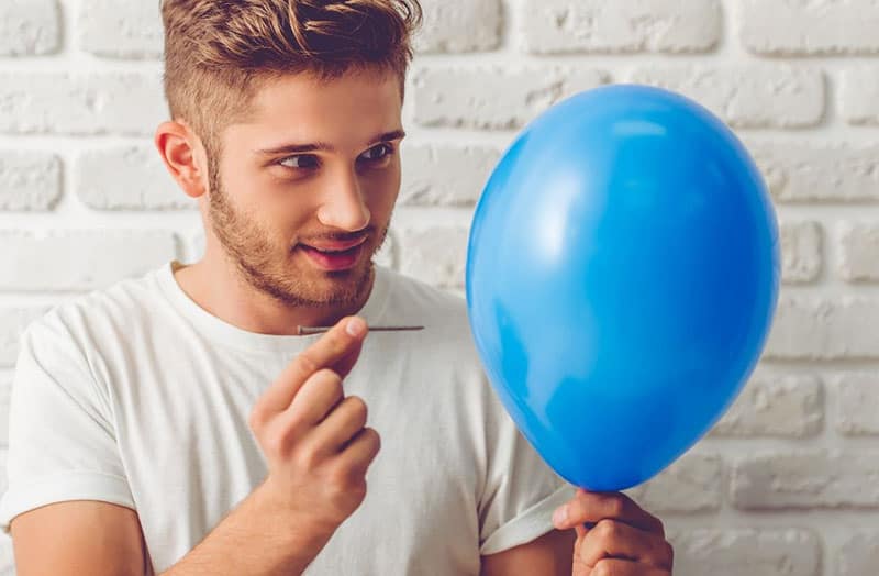 guy about to burst a blue balloon with a needle near a white brick wall