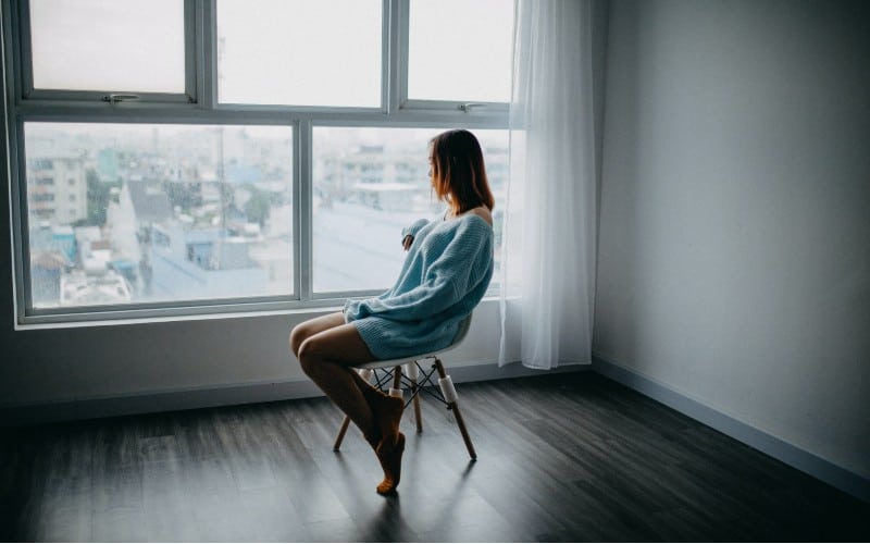 Sad lonely woman sitting on brown chair beside window in empy room during daytime