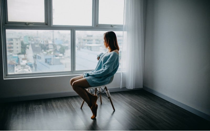 Triste mujer solitaria sentada en una silla marrón junto a la ventana en una habitación vacía durante el día.