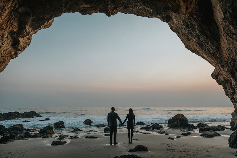 man and woman hold hands at beach with rocks