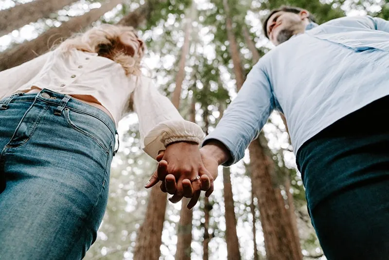 man and woman holding each others hands while standing beside the trees