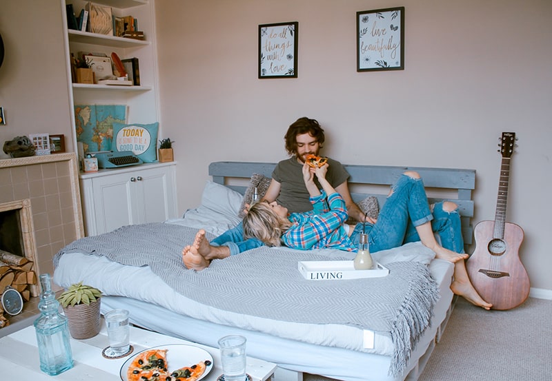 man and woman lying on bed while eating pizza