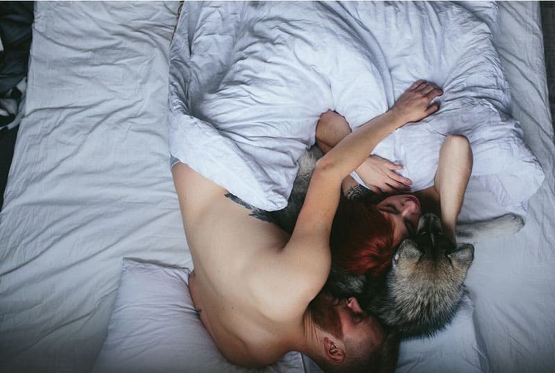 man and woman sleeping with man's hand over the woman and a dog in between them in white bed and linens