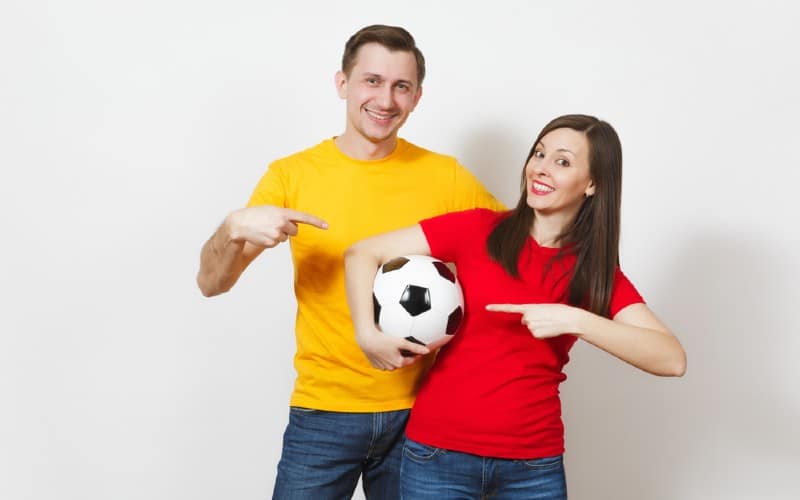 Hombre feliz con camiseta amarilla y mujer con camiseta roja sosteniendo un balón de fútbol.