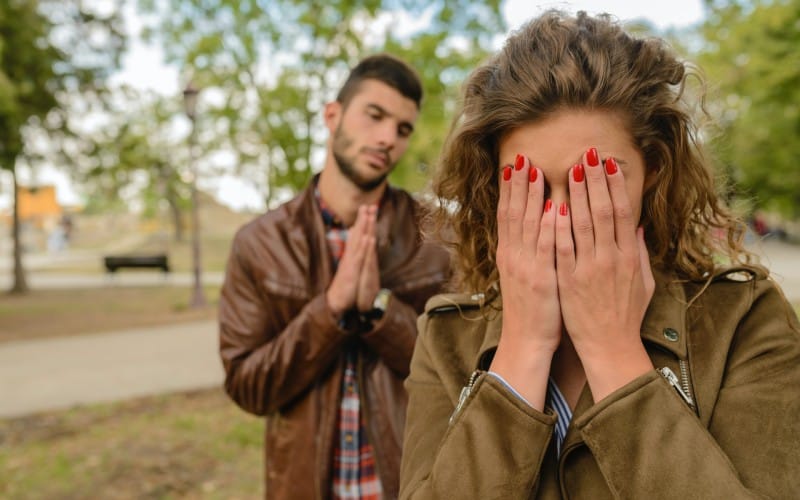 Man in brown jacket asking for forgiveness beside woman outdoors during daytime