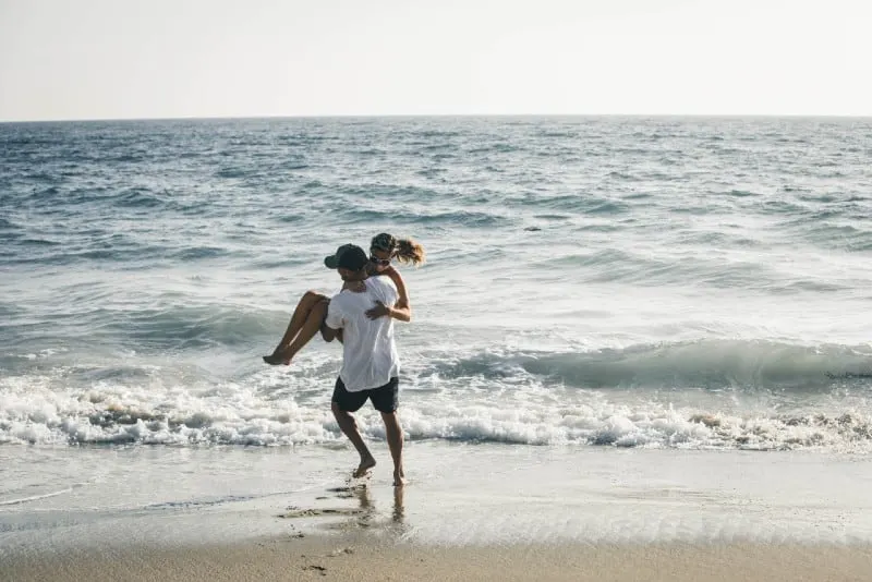 man in white shirt carrying woman on beach