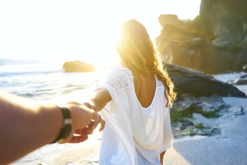 homem a segurar a mão de uma mulher junto ao mar, de frente para o pôr do sol