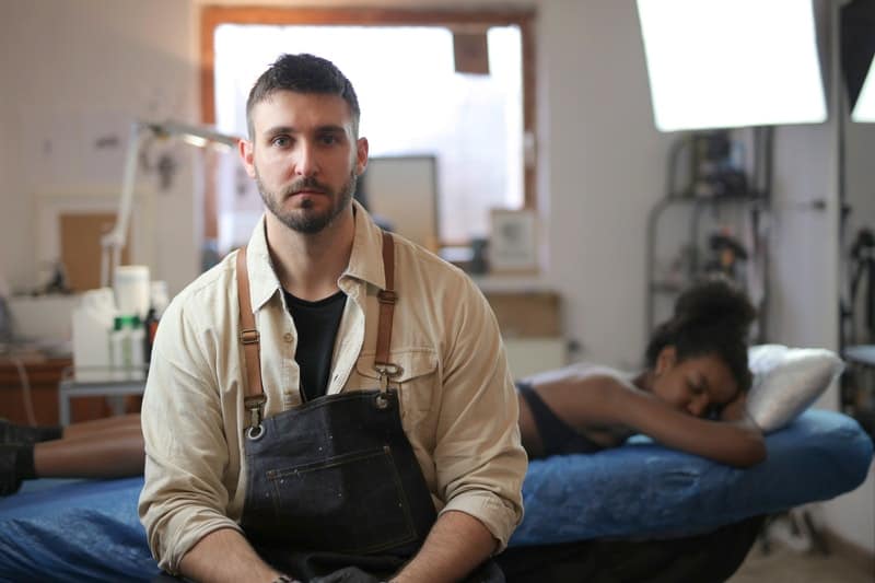 man in beige and apron sits while a woman sleeps at the back 