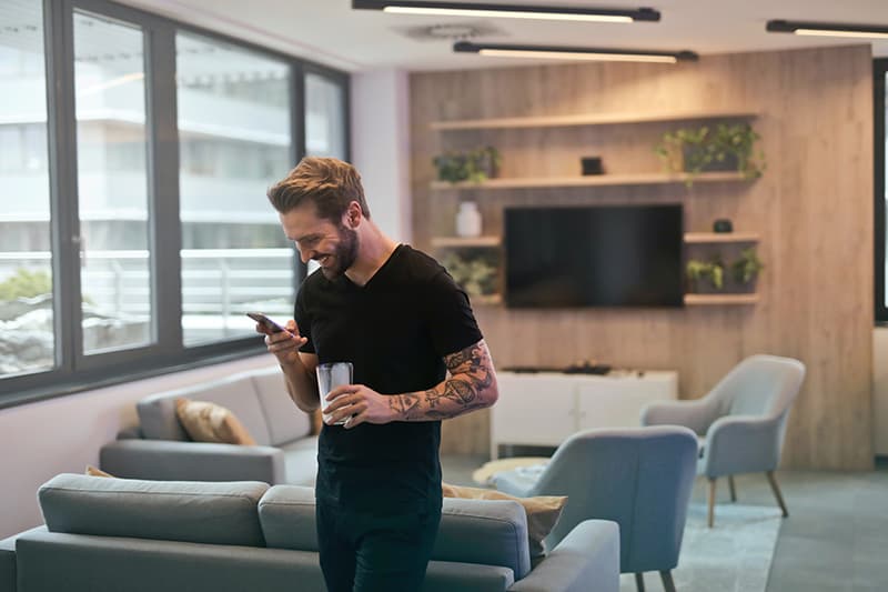 hombre con camisa negra de pie mientras mira un smartphone con un vaso en la mano izquierda