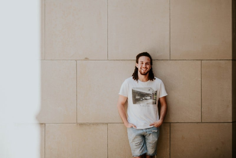 hombre con camisa blanca estampada apoyado en la pared y sonriendo