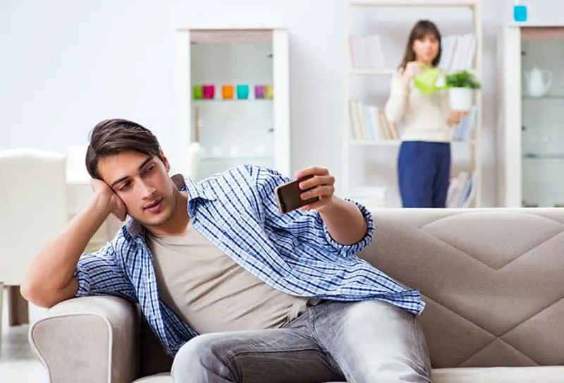 man leans on couch looking at his cellphone with a woman standing at a distance