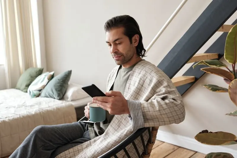 man looking at his cellphone while having his coffee and on bath robe sitting