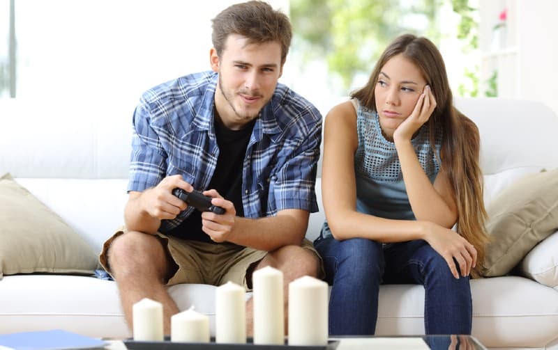 Man playing video games sitting on a couch near bored woman