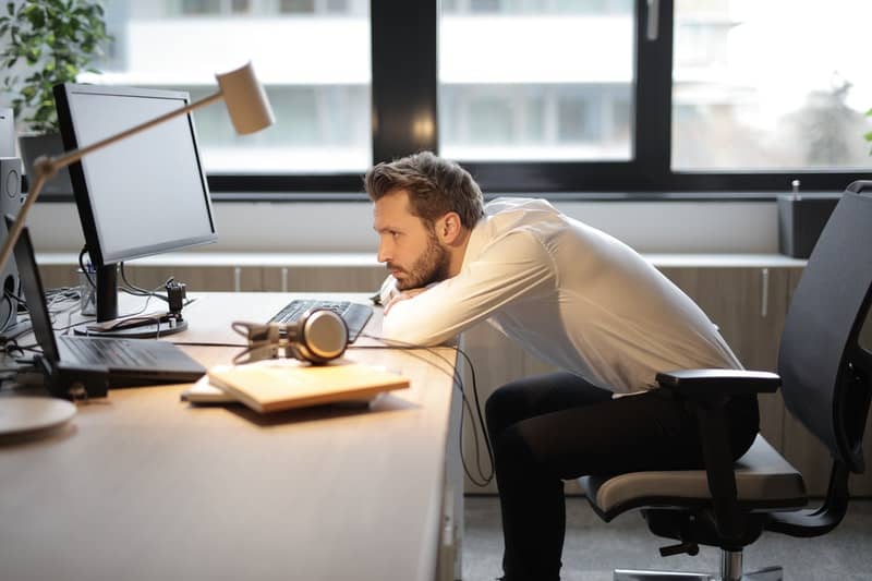 man sits on chair leaning his head on the table looking at the computer monitor